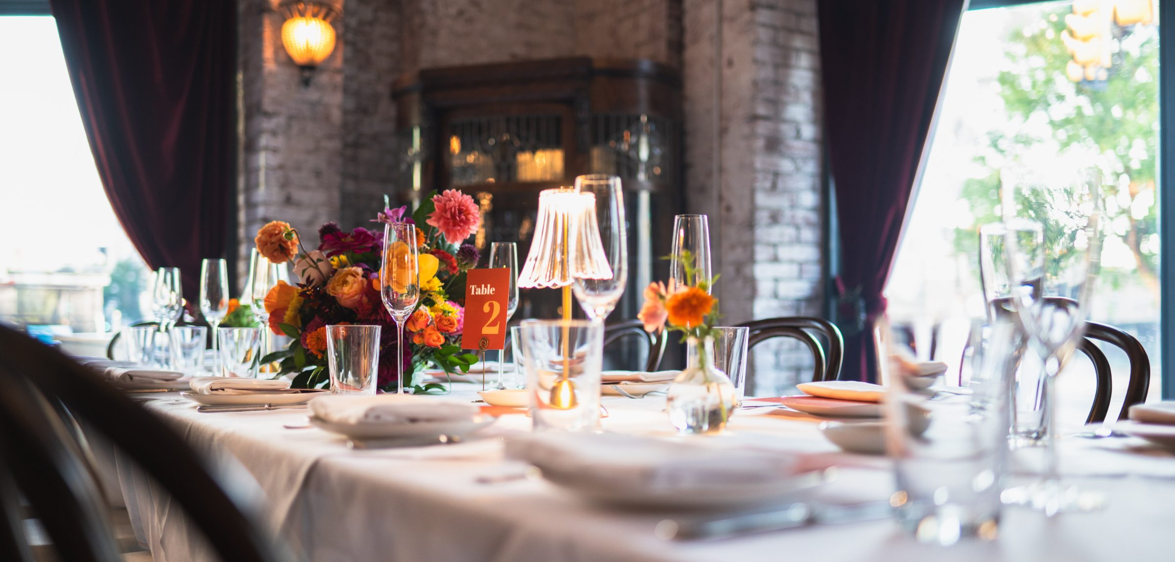 A dressed table under a shaded lamp — glassware, linen and autumn flowers against exposed brick and deep red drapes.