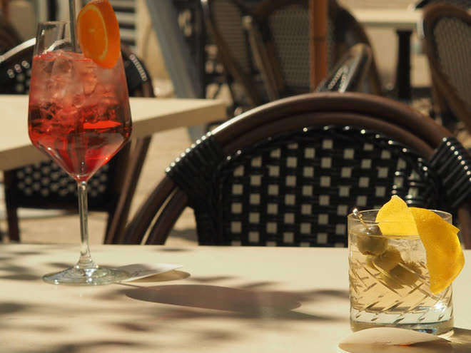 A spritz and a lemon-peel cocktail on a bistro table in dappled sunlight.