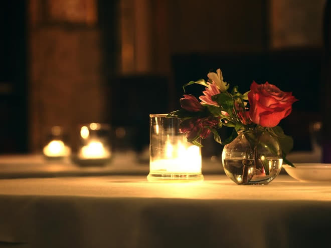 A lit candle and a small vase of roses on a dark table.
