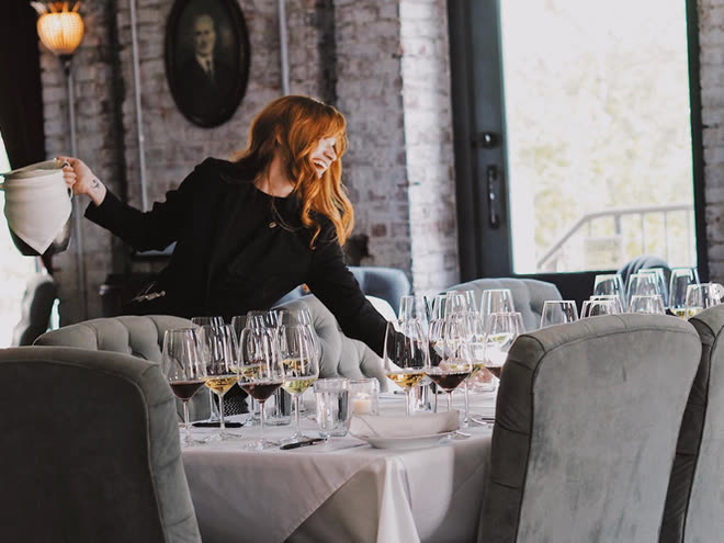 A woman in black setting wine glasses along a dressed table by a bright window.
