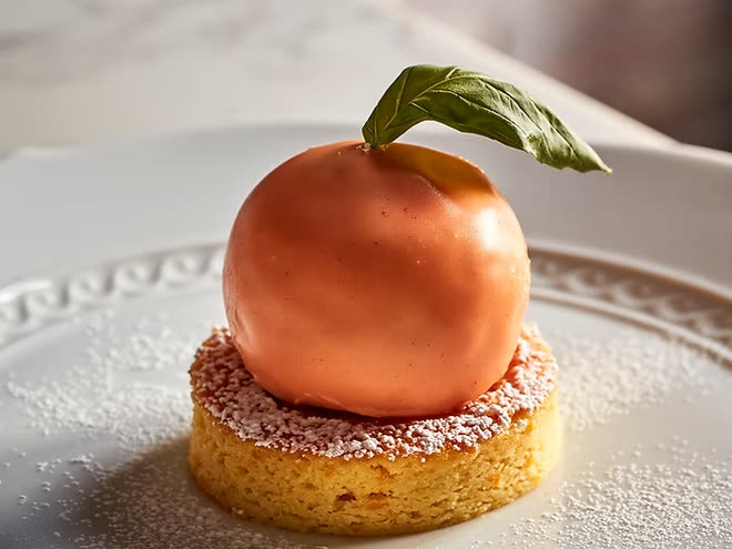 A peach-shaped dessert finished with a basil leaf, on a sponge base dusted with icing sugar.