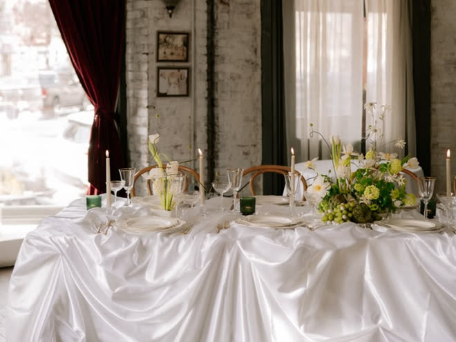 A long table dressed in white satin with candles and white flowers by a window.