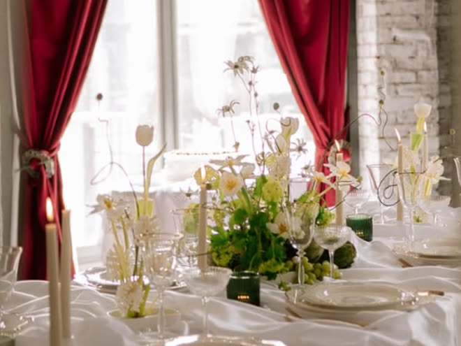 A dressed table in white linen with spring flowers and lit candles against red drapes.