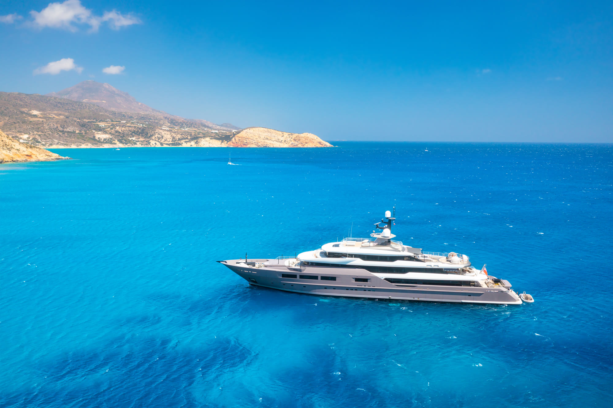 A yacht anchored in clear Aegean water off a rocky shore.